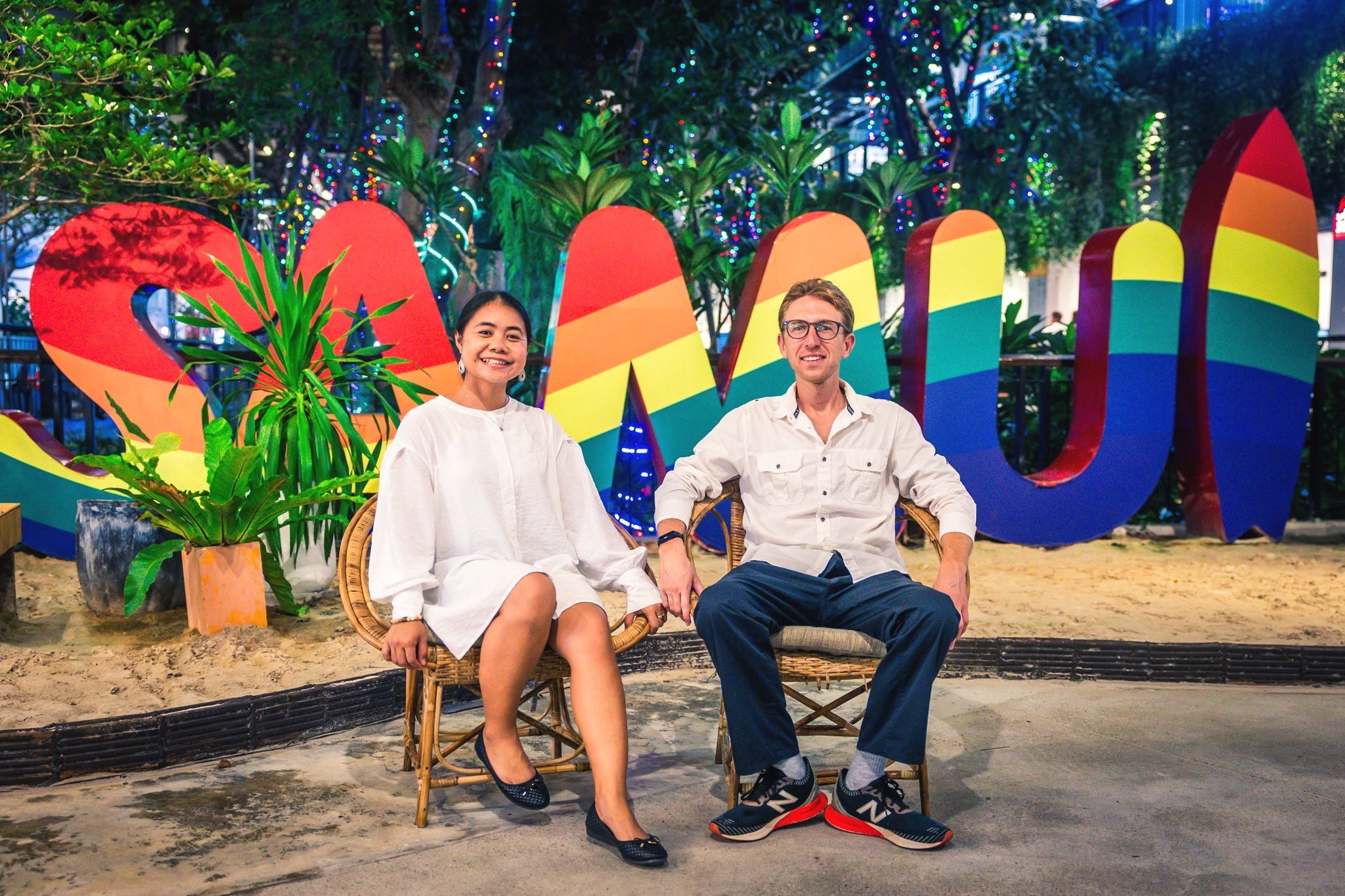 A man and a woman posing on seats in front of a colorful Samui sign.